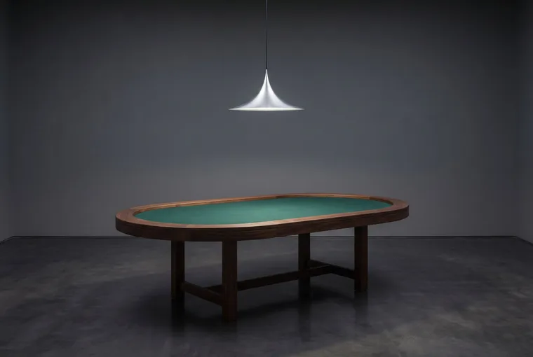 Une table de cartes ovale, vide, à tapis vert, sous une suspension en aluminium.